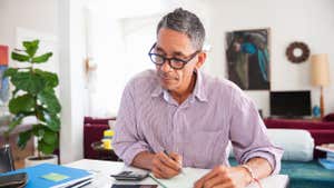A man with gray hair sits at a desk, using a calculator and a pen and paper.