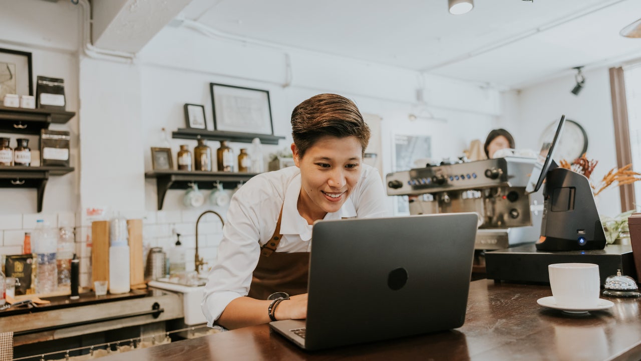 A smiling coffee shop owner leans over the counter, working on a laptop.